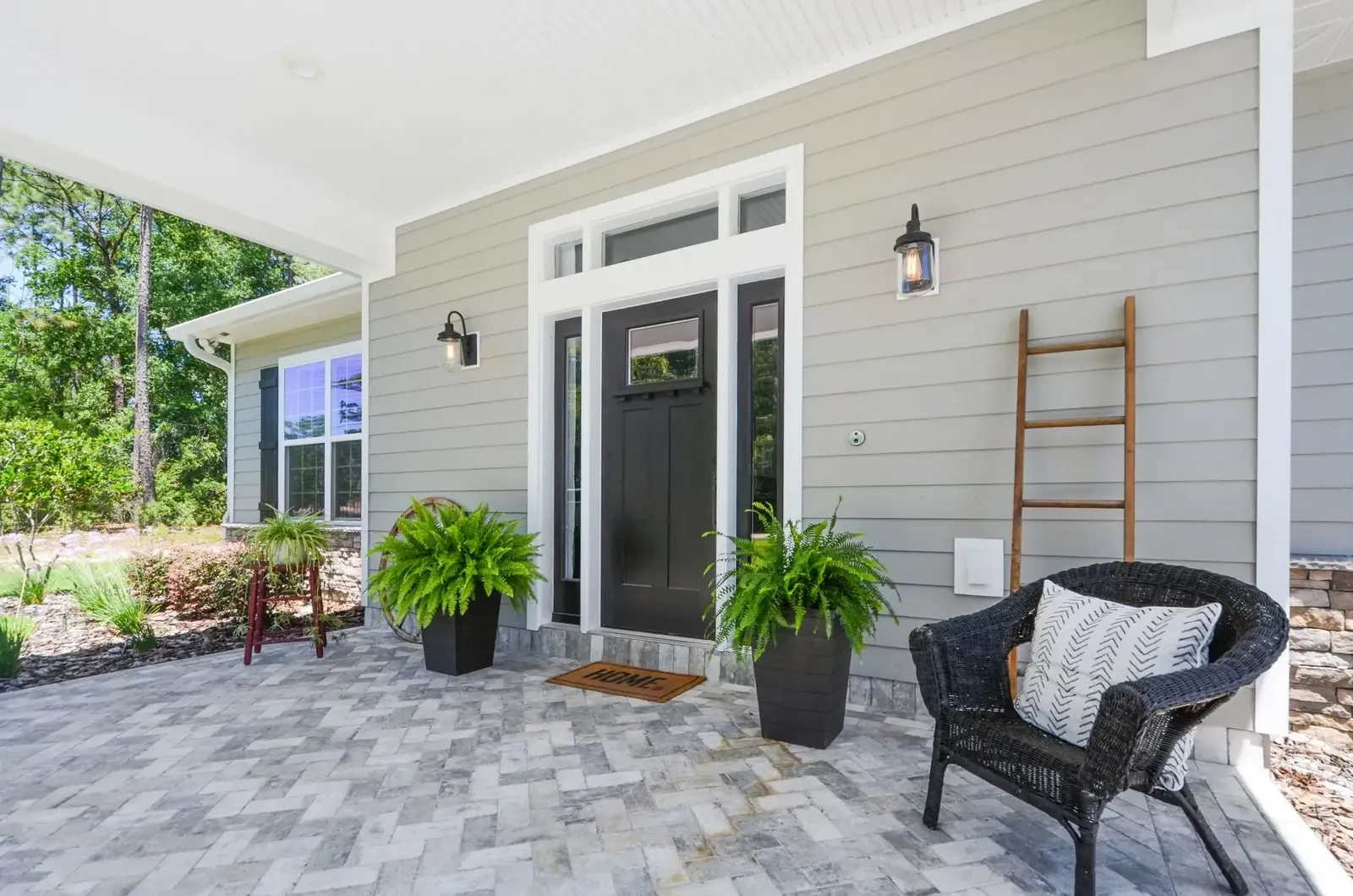 Covered front porch with paver flooring, a black front door and a wicker chair on a custom home
