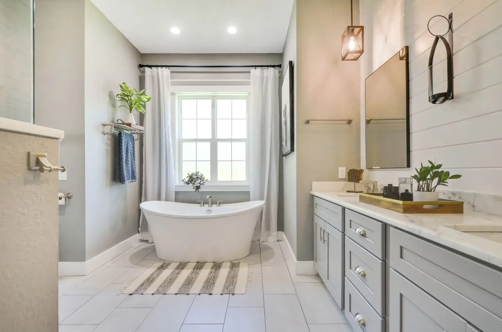 Primary bathroom with a freestanding tub, gray vanity cabinets and a window with white curtains