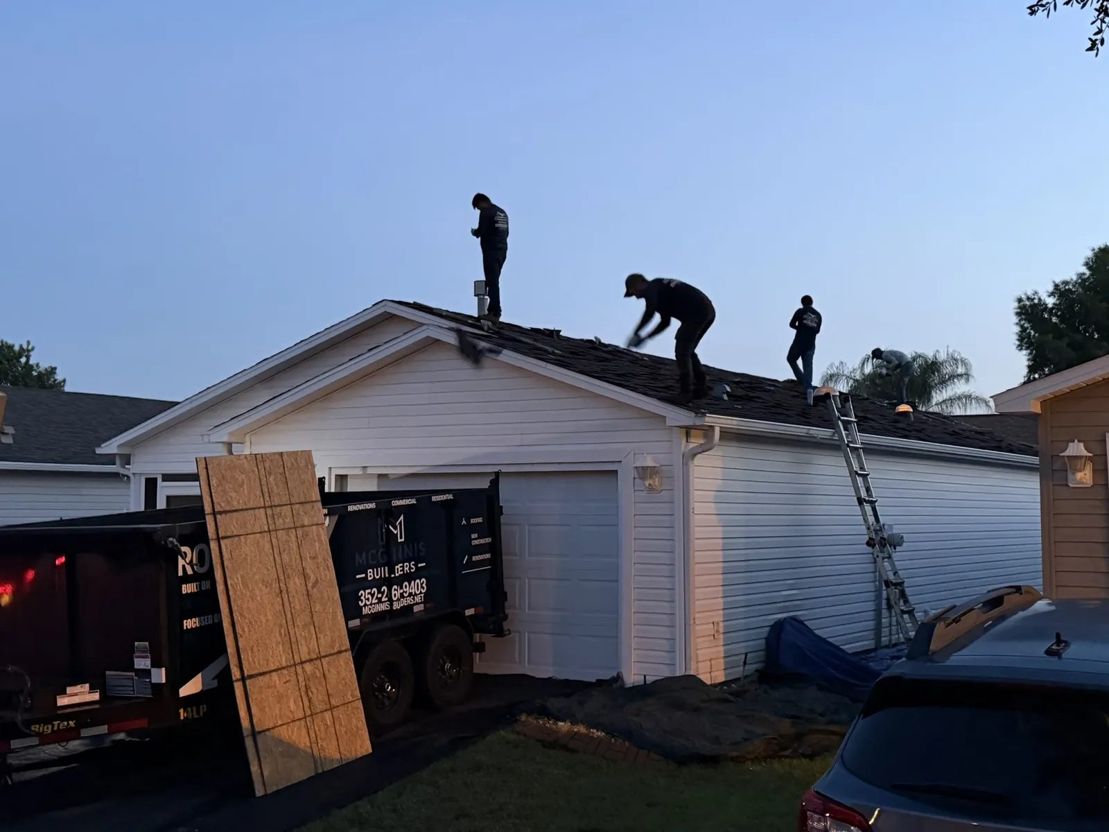 McGinnis Builders crew tearing off an old roof at sunrise, with a roofing trailer in the driveway