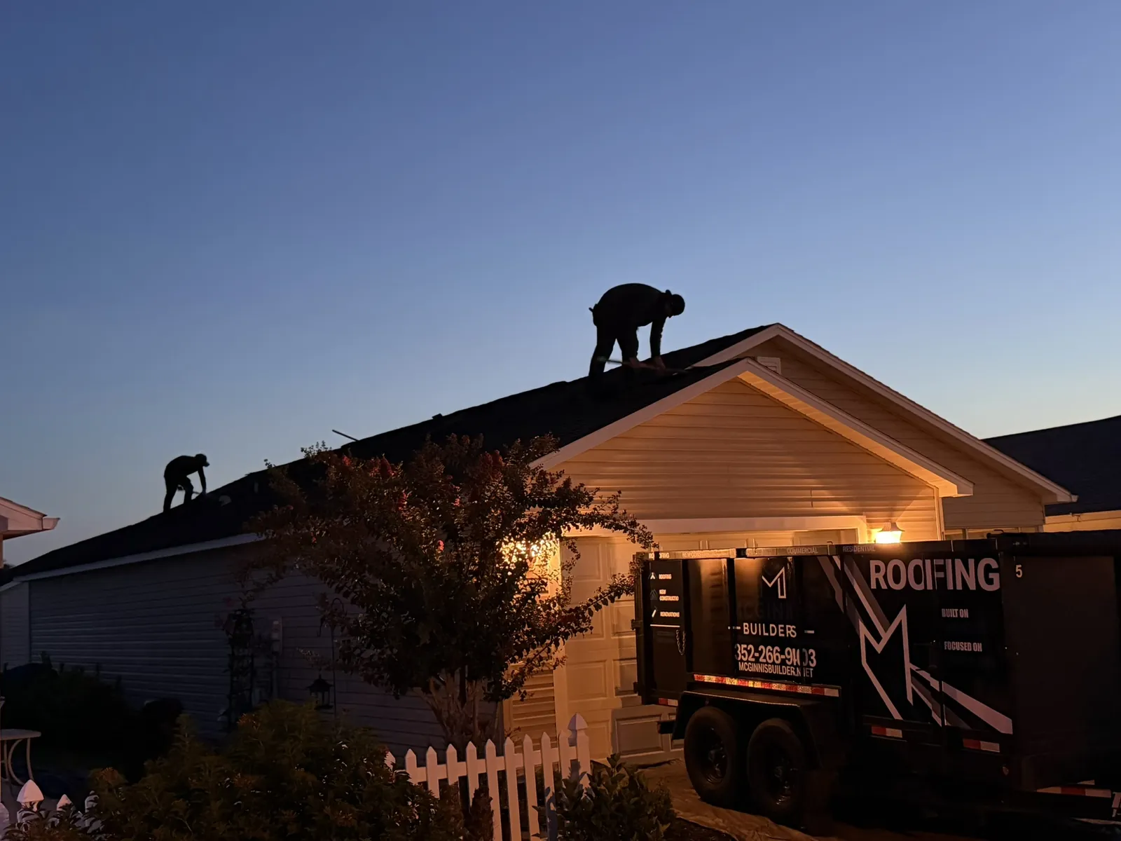 Roofer working at dawn beside a McGinnis Builders roofing trailer