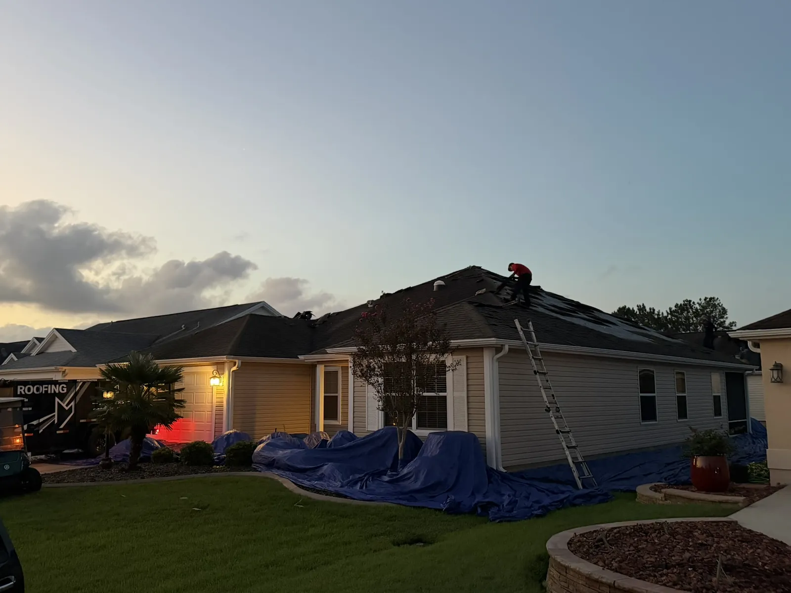 Roofer installing new shingles at dusk while tarps protect the landscaping